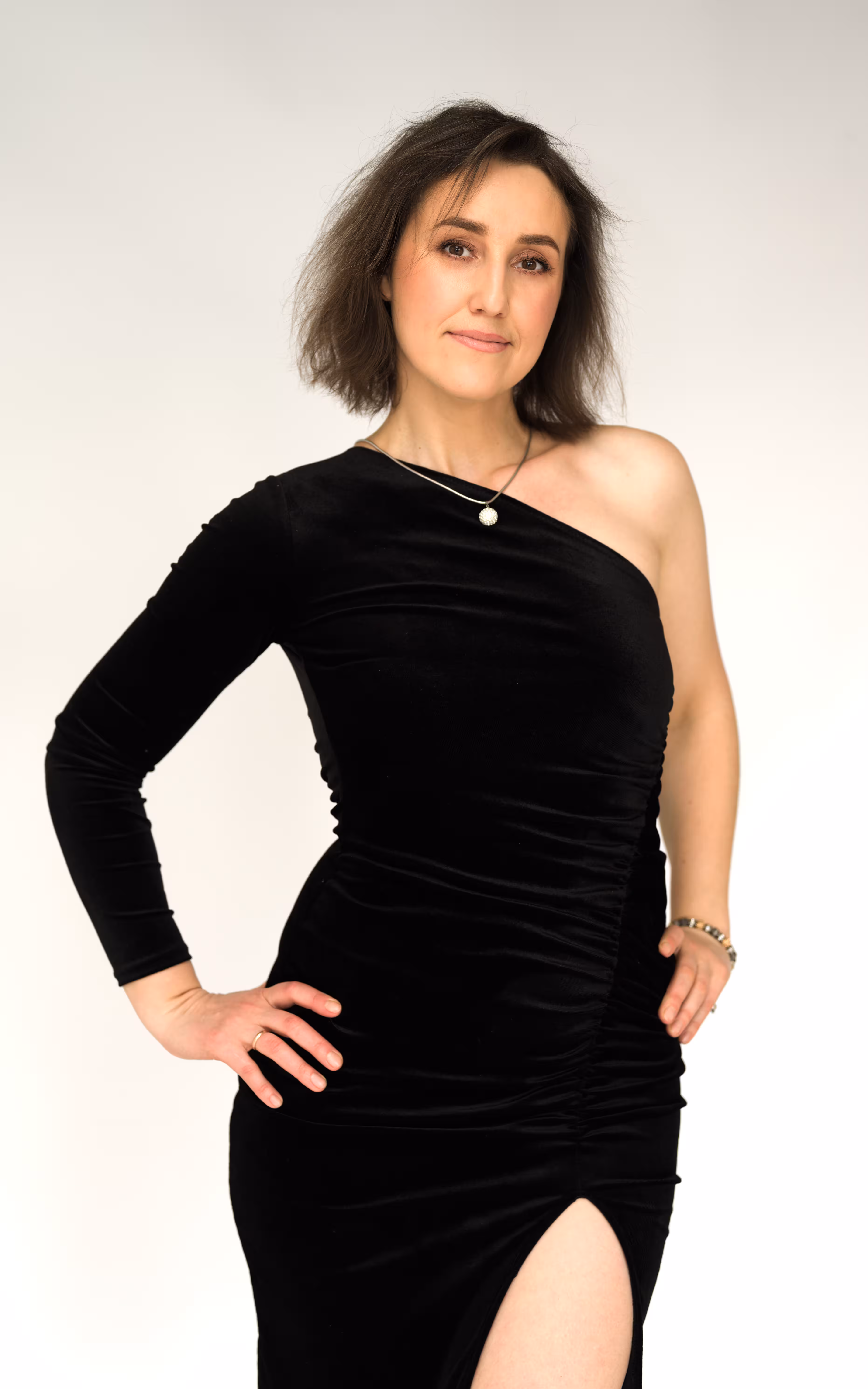 A medium shot of a woman with brown hair posing against a light gray background. She is wearing a form-fitting black one-shoulder velvet dress with a side slit. She stands with both hands on her hips, looking directly at the camera with a subtle, confident smile.