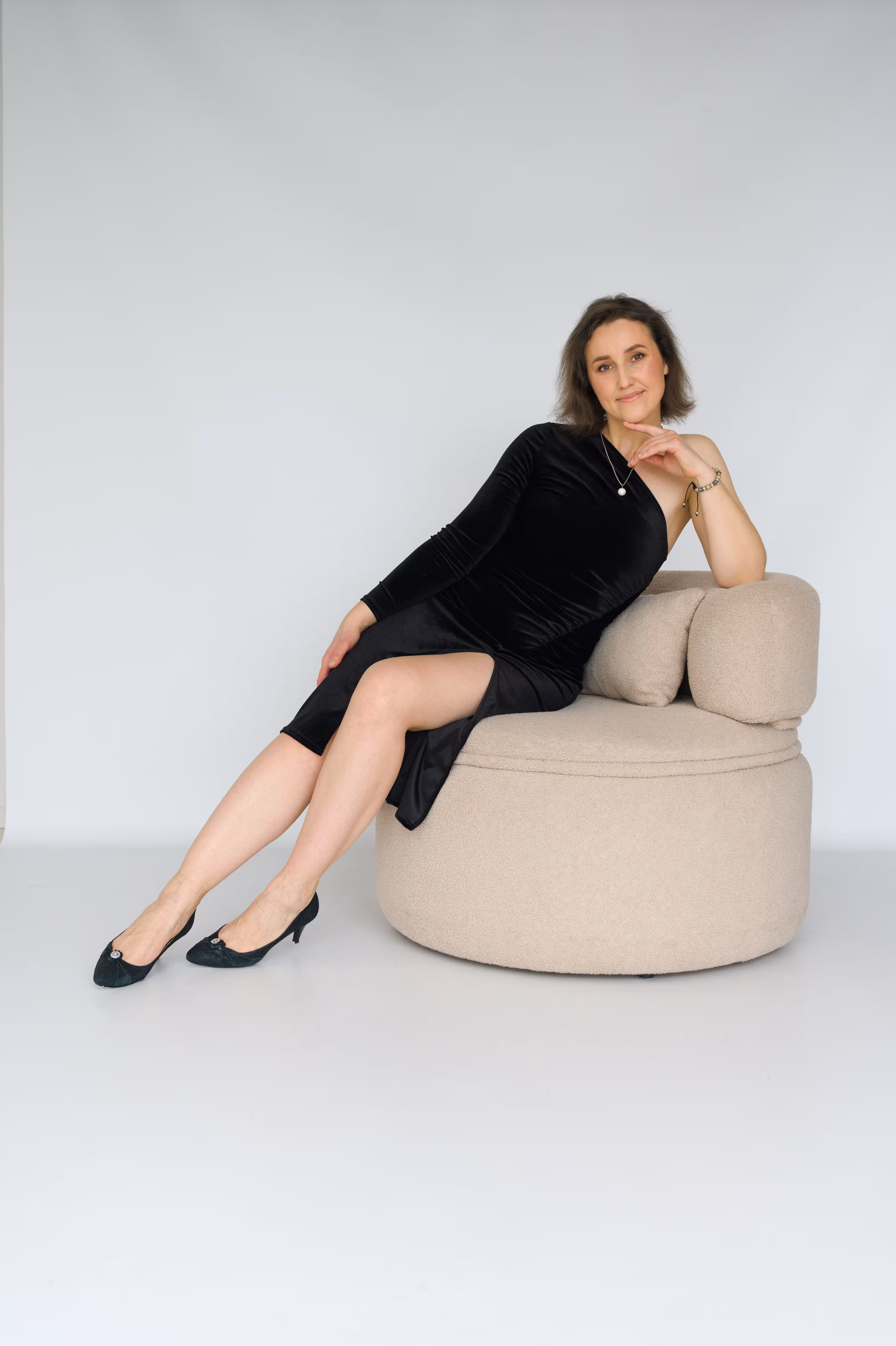 A woman with brown hair sits elegantly on a round, beige textured armchair against a white background. She is wearing a black one-shoulder velvet midi dress with a high leg slit and black low-heeled shoes. She is posing with her hand gently resting under her chin and a soft smile.